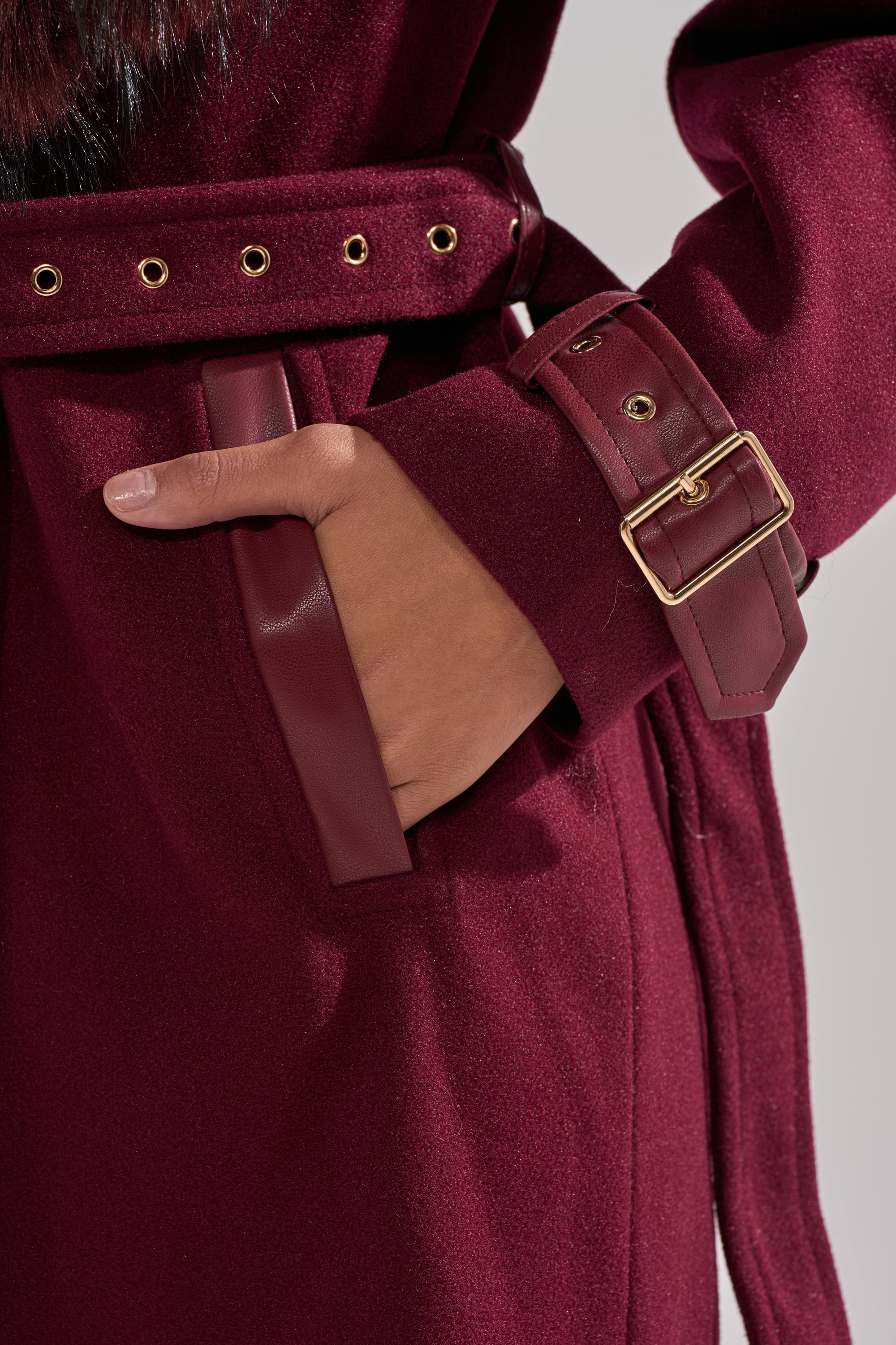 A close-up of a hand in the pocket of the CATALINA FUR COLLAR TRENCH, a burgundy faux fur coat with leather trim, a matching belt featuring gold eyelets and buckle. 14