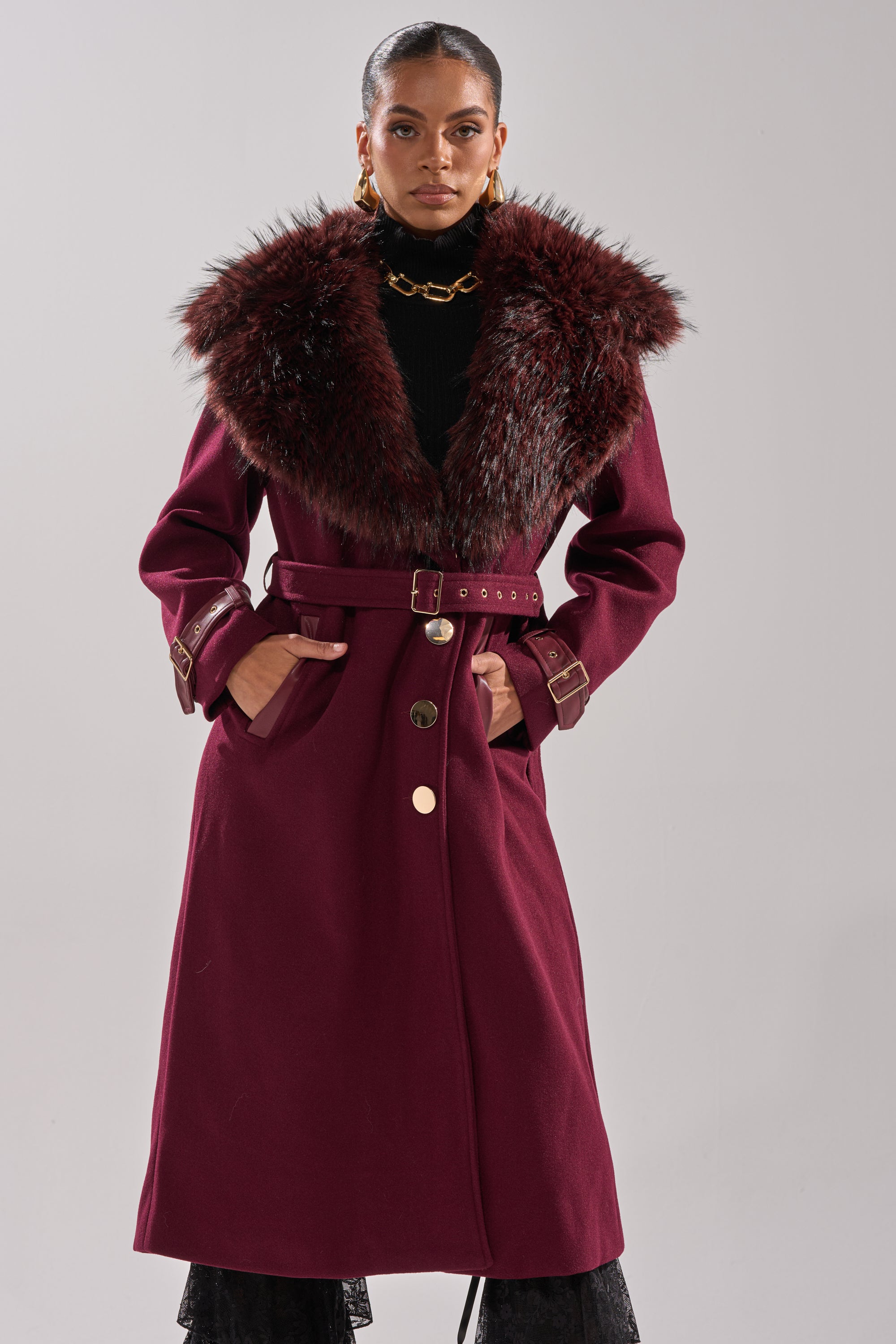 A model wears the CATALINA FUR COLLAR TRENCH—a long burgundy coat with a wide dark faux fur collar and matching belt—styled with gold earrings and a chunky necklace against a plain light gray background. 1