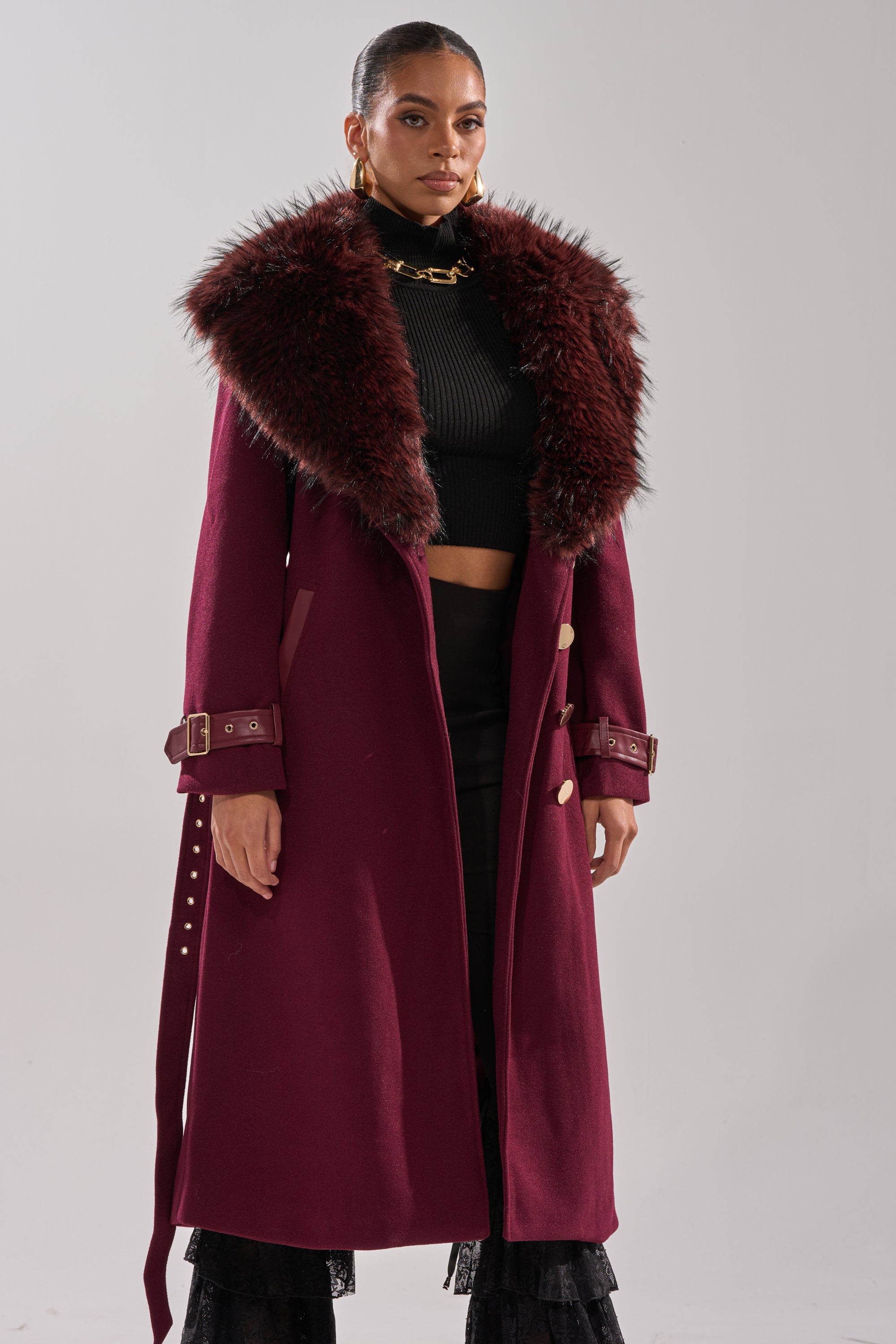 A woman stands against a plain light background wearing the CATALINA FUR COLLAR TRENCH, styled with a black cropped turtleneck, black pants, and gold hoop earrings. 2