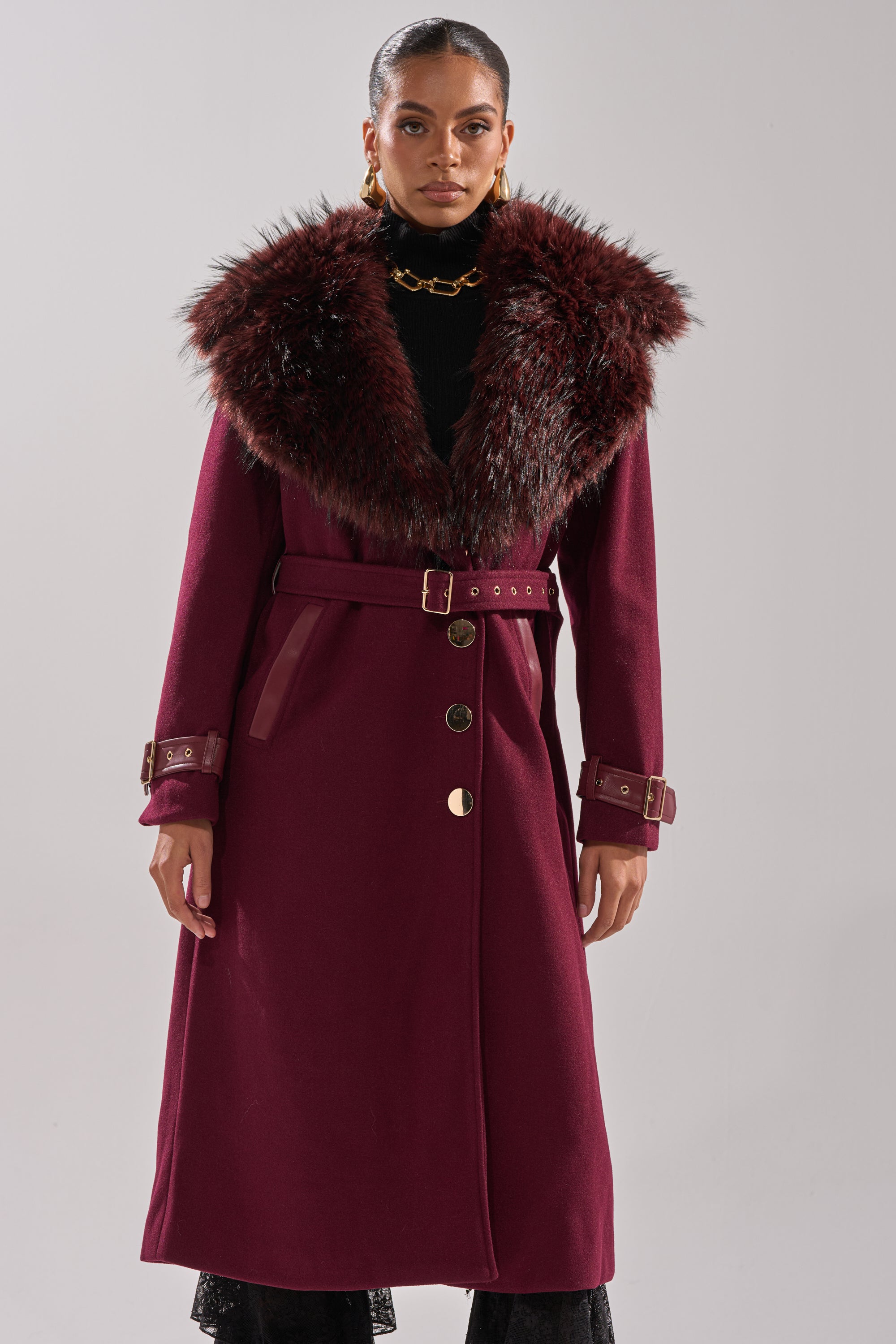 Wearing the CATALINA FUR COLLAR TRENCH, which features a longline cut, wide fluffy fur collar, gold buttons and buckles, and a belted waist, a person stands against a plain light background. 3
