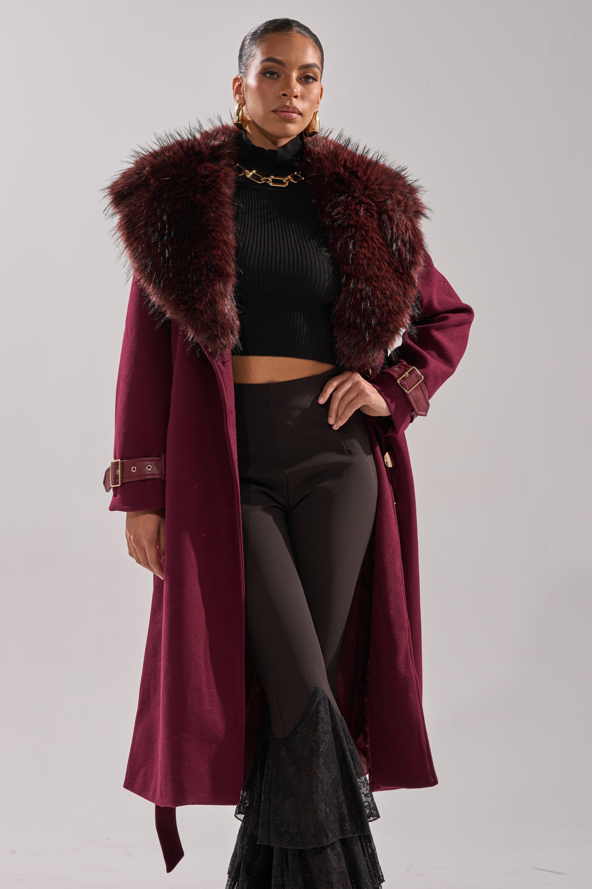 A woman poses confidently in the CATALINA FUR COLLAR TRENCH, styled with a black cropped turtleneck, black high-waisted lace flare pants, gold earrings, and a chunky gold chain necklace. 4