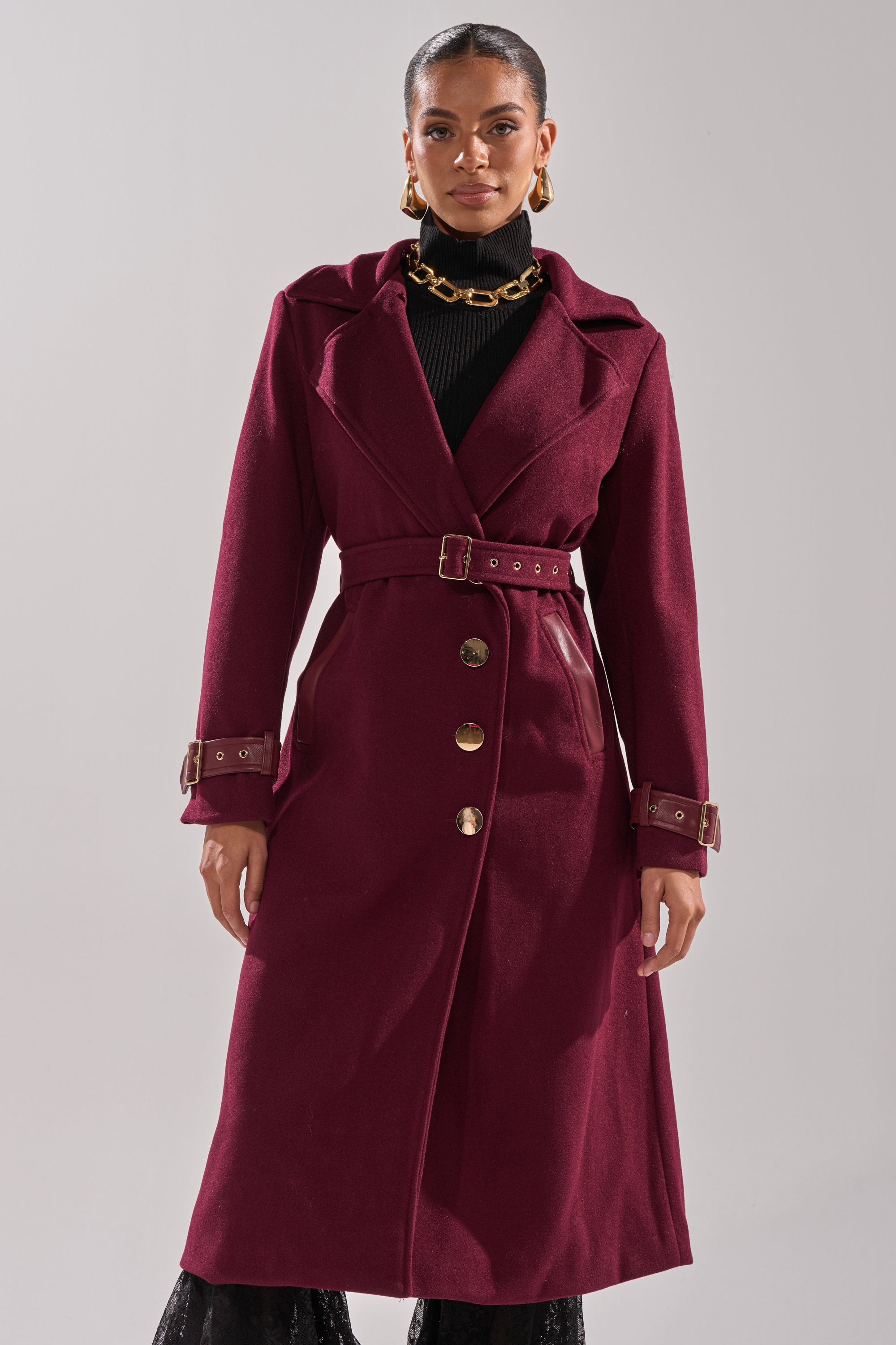 A woman wears the CATALINA FUR COLLAR TRENCH, a long burgundy belted coat with gold buttons and buckled cuffs, over a black turtleneck. She completes her look with gold hoop earrings and a chunky necklace against a light background. 5