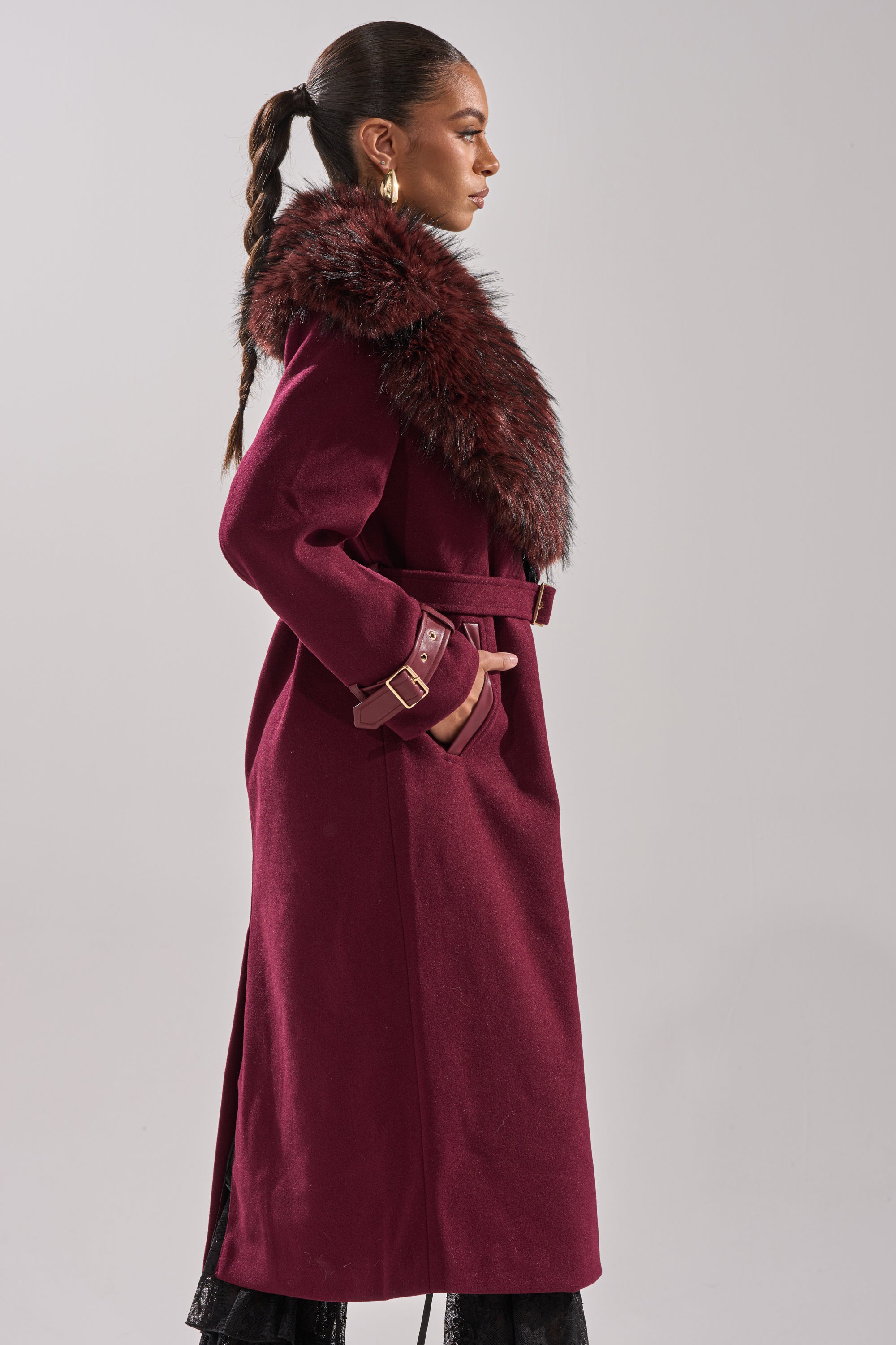 A woman stands in profile wearing the CATALINA FUR COLLAR TRENCH, a longline jacket with a burgundy faux fur collar, belted waist and cuffs. She accessorizes with gold hoop earrings and has a sleek, braided ponytail. 6