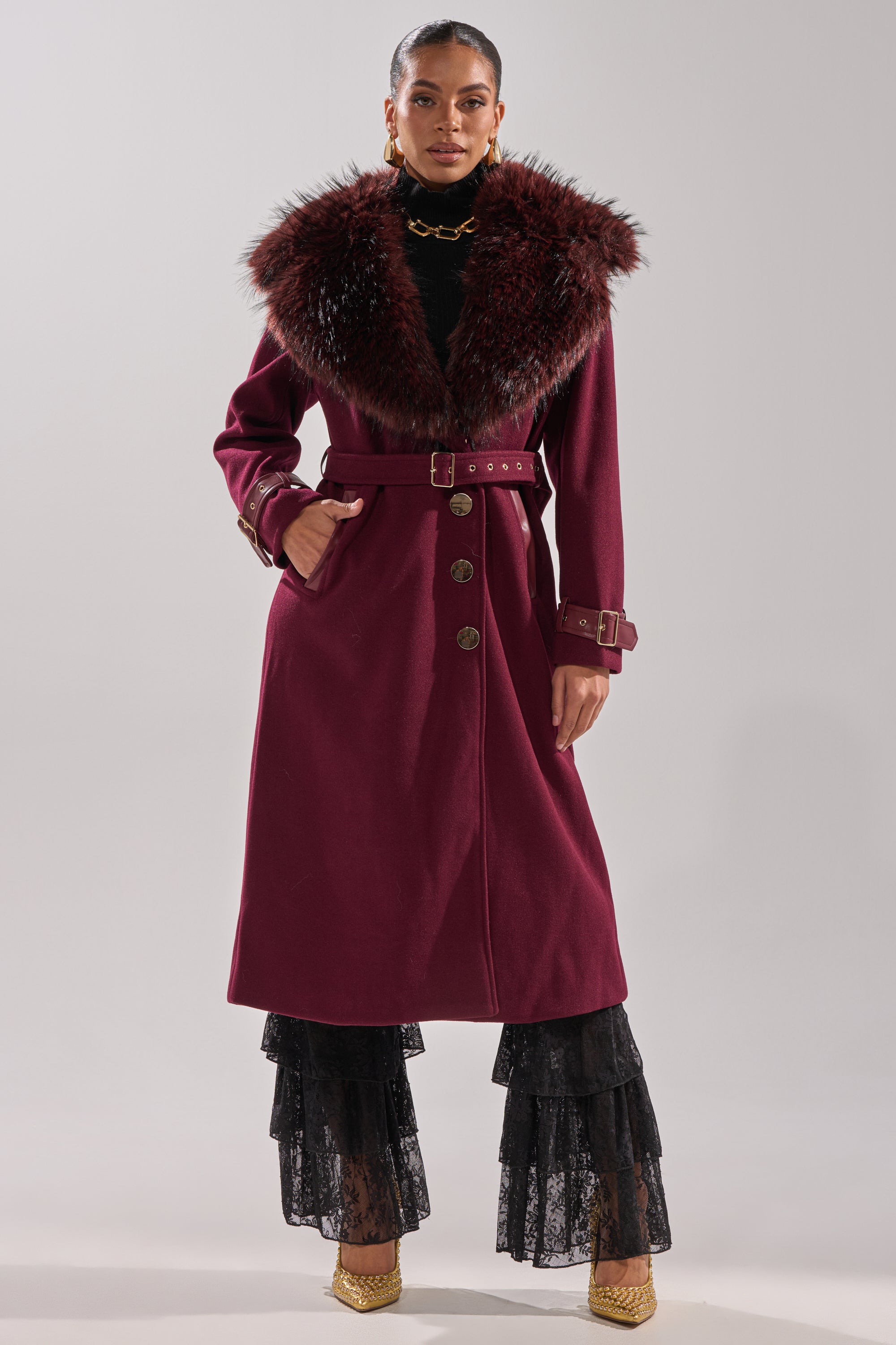Confidently dressed, she wears the CATALINA FUR COLLAR TRENCH—a longline coat with a wide fur collar, gold buttons, and matching belt—styled with black lace tiered pants, gold heels, hoop earrings, and a chunky necklace. 8