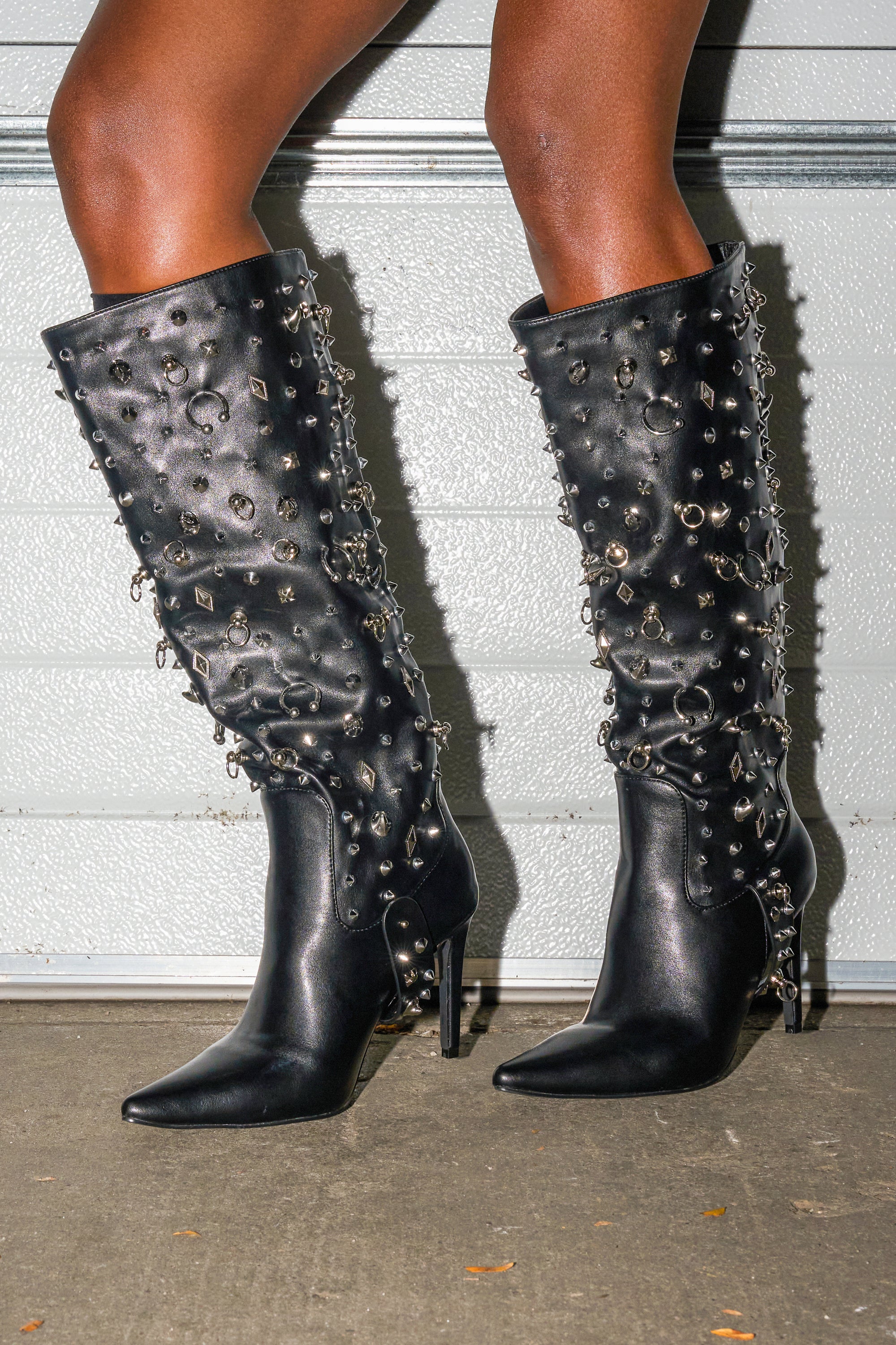 Someone wearing the AZALEA WANG AKRISTA BLACK HARDWARE EMBELLISHED STILETTO KNEE HIGH BOOT with silver studs, rings, and chains stands on concrete in front of a light-colored garage door.