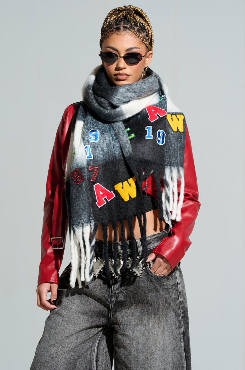 A person wearing a red leather jacket and black sunglasses stands confidently with their hands in their pockets. Featuring a braided hairstyle, they accessorize with the PATCHED UP PLAID SCARF as a winter accessory and complete the look with high-waisted gray jeans.