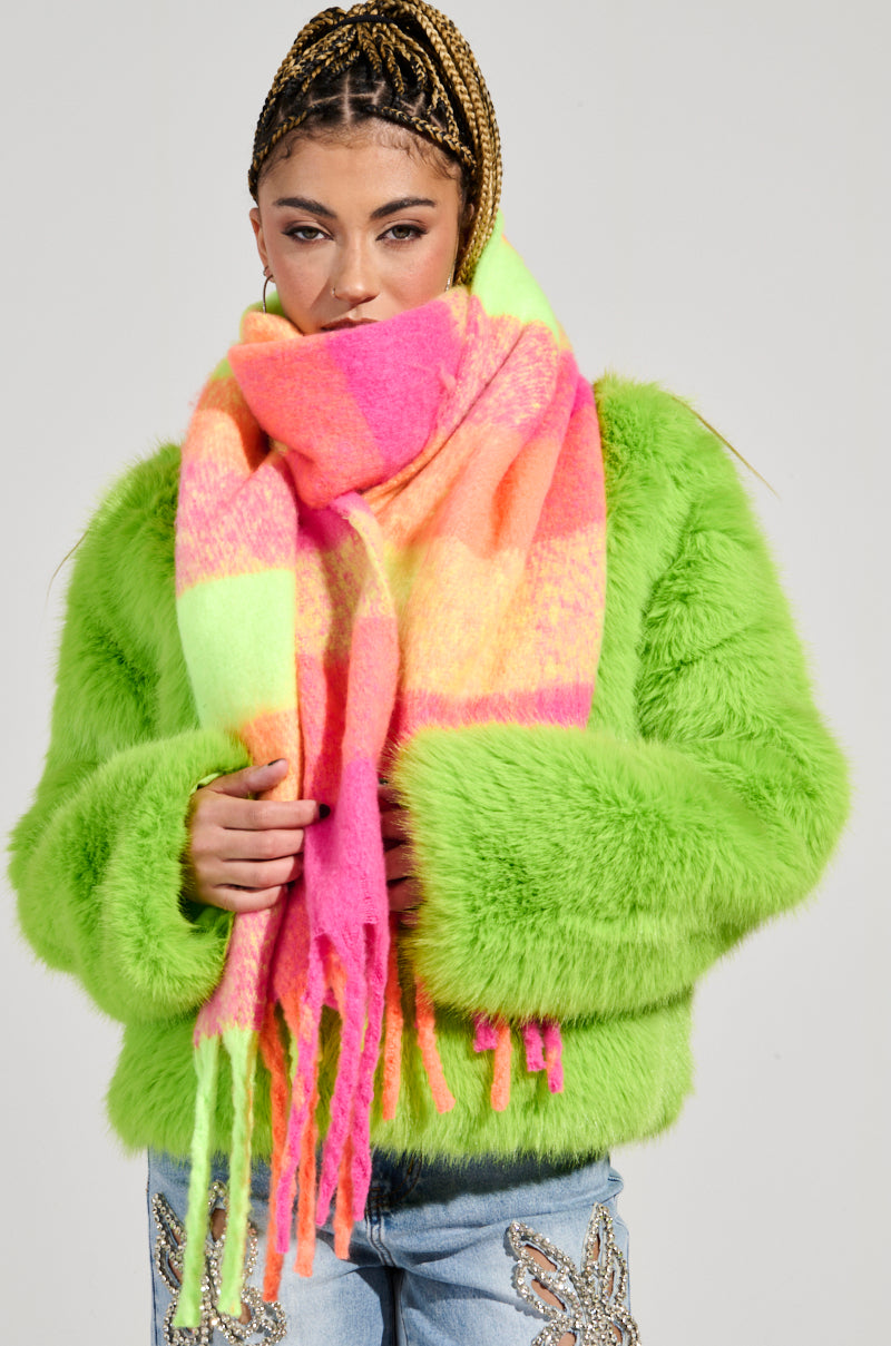 A person wearing a vibrant green faux fur jacket and an oversized UNDER THE SUN PLAID SCARF featuring stripes in pink, yellow, and orange. Their braided hair complements their jeans adorned with decorative designs and fringe trim detailing against a plain, light-colored background.