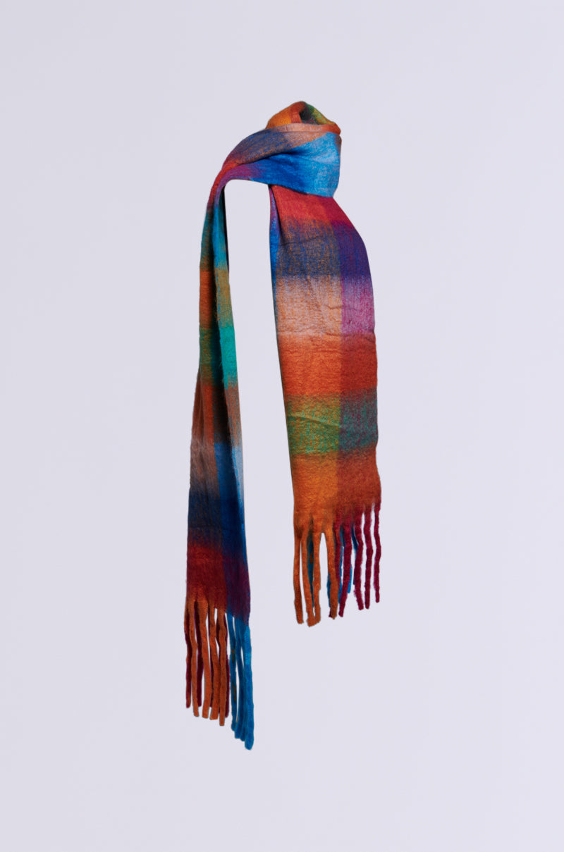 The BY THE FIRE BLANKET SCARF from the AKIRA Label boasts a dynamic plaid pattern in lively hues of blue, orange, red, and green. Its fringed ends create a striking contrast against a simple gray backdrop.
