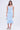 A woman with long brown hair in loose waves stands against a plain background wearing the LUNA BANDAGE MIDI SKIRT in light blue, featuring a stretchy bandage knit and flared hem. She holds a silver handbag and wears silver heeled sandals.