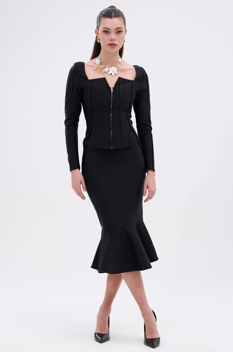 A woman stands against a plain background wearing the JESSIE BANDAGE MIDI SKIRT, styled with a fitted black top, statement necklace, sleek hair, and black high heels for a chic and sophisticated look. 0