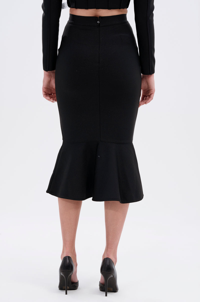 A person is shown from the back against a plain light background, wearing the JESSIE BANDAGE MIDI SKIRT—a fitted black bandage skirt with a flared peplum hem—paired with black high heels. 3