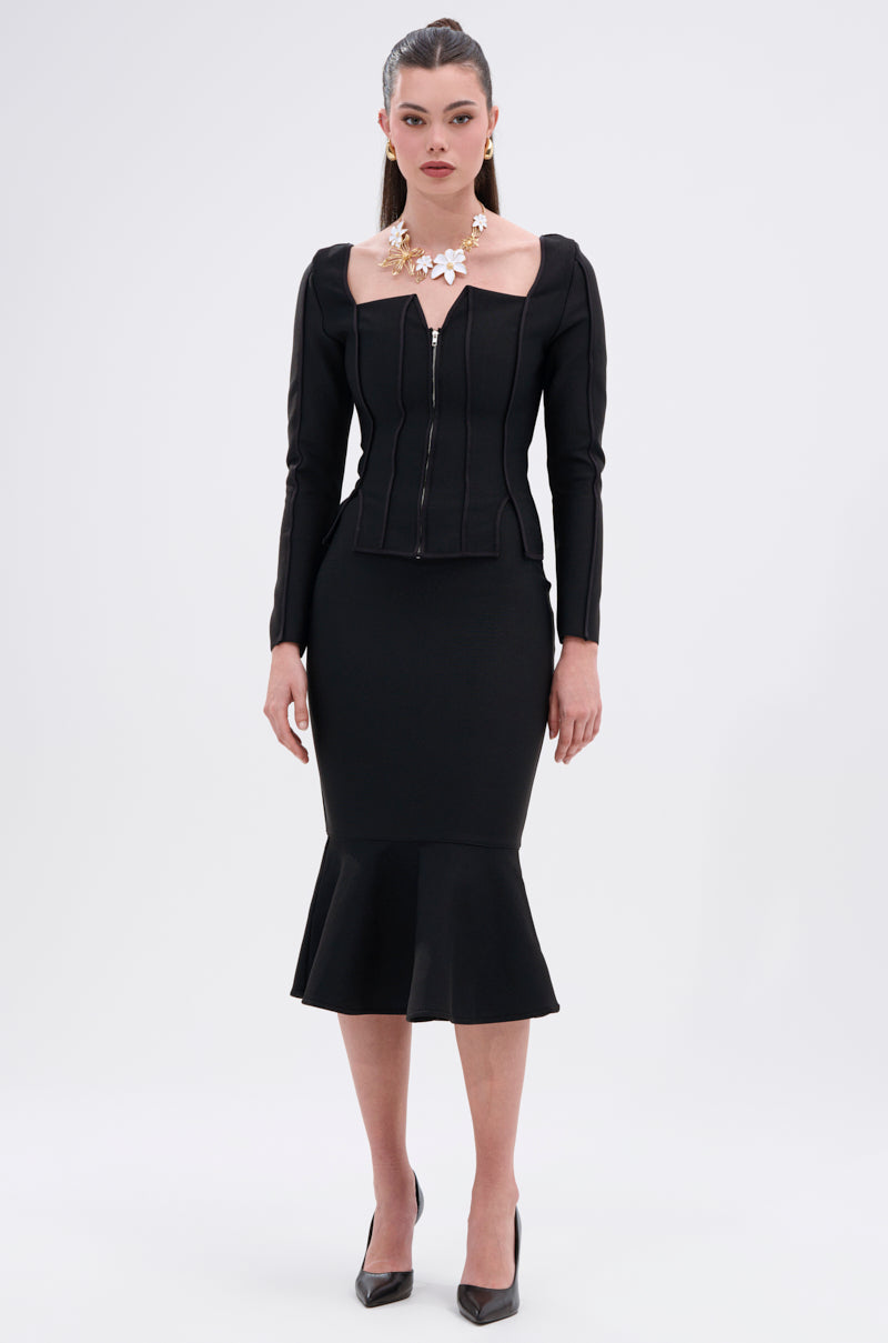 A woman poses against a plain background wearing the JESSIE BANDAGE MIDI SKIRT, styled with black heels and a chunky gold necklace featuring white accents. The outfit highlights a fitted silhouette and sophisticated flair. 5