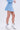 A woman stands against a plain light background wearing the ICY BLUE SPARKLE MINI SKIRT with a matching skater silhouette top, white frilly ankle socks featuring black bows, and silver high-heeled sandals.