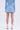 A person stands facing away against a plain white background, wearing a light blue long-sleeve top, the ICY BLUE SPARKLE MINI SKIRT with a skater silhouette, white socks, and silver high-heeled shoes.