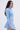 A woman with long wavy brown hair stands in profile against a plain light background, wearing the ICY BLUE SPARKLE CARDIGAN and a matching pleated skirt.