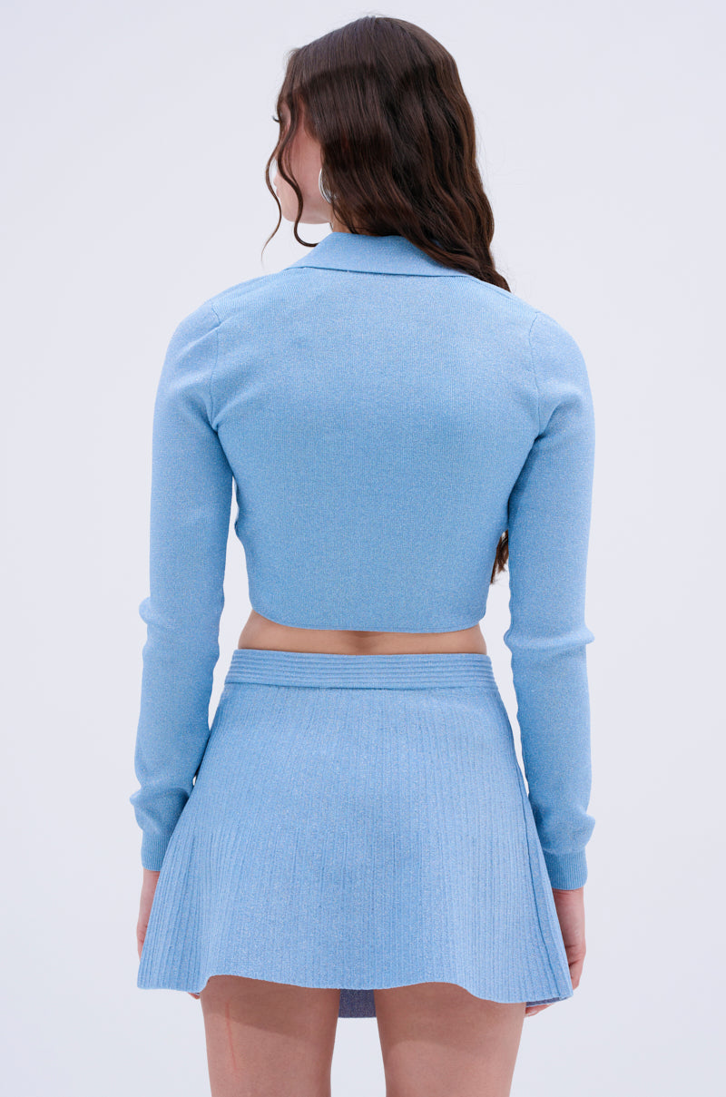 A woman with long, wavy brown hair is shown from the back wearing the ICY BLUE SPARKLE CARDIGAN and a matching pleated mini skirt. The background is plain and light-colored.
