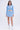 A woman faces forward in the ICY BLUE SPARKLE CARDIGAN and a matching mini skirt, paired with white socks featuring black bows and clear-heeled sandals. She has long wavy hair, a neutral expression, and stands before a plain light backdrop.