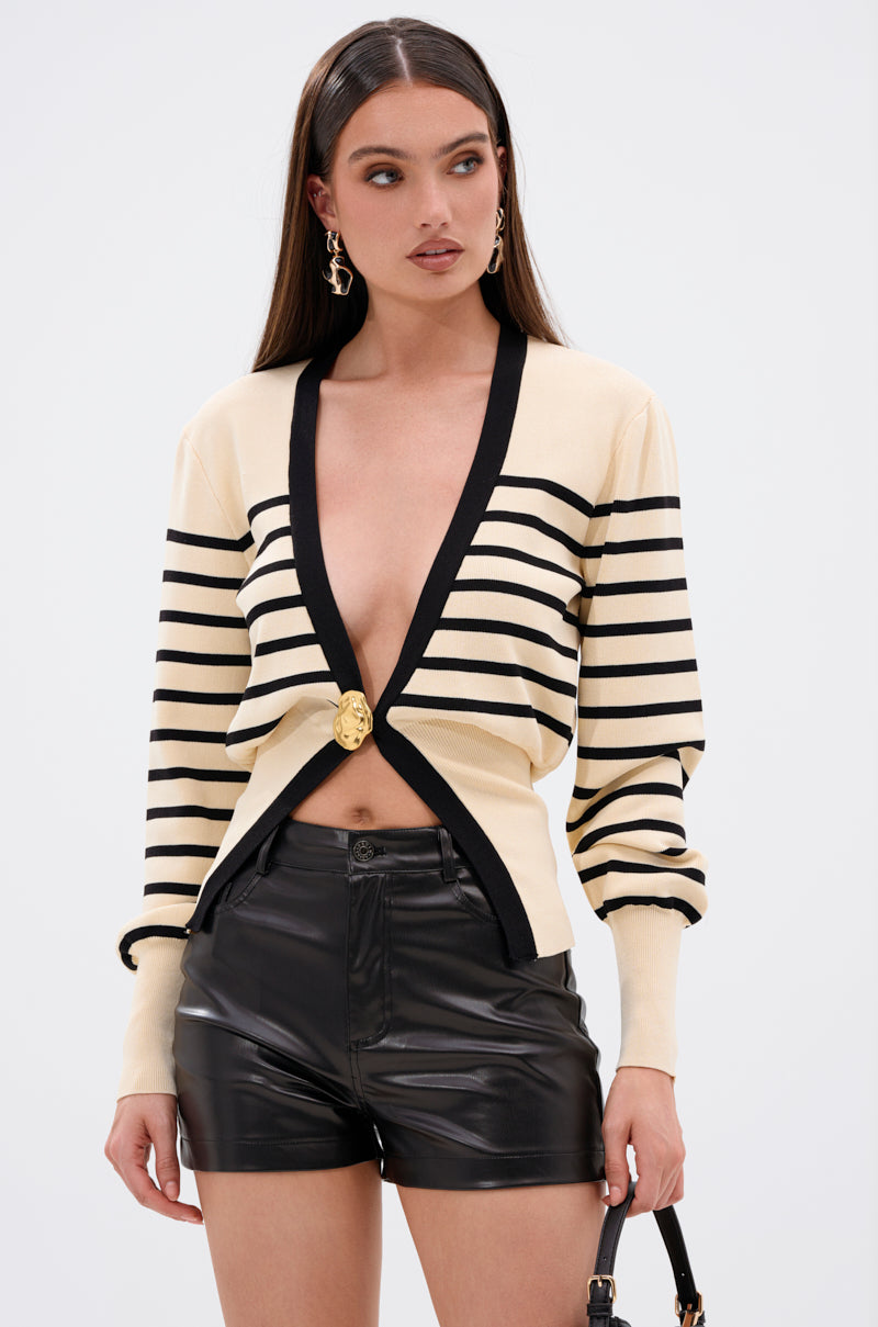 A woman wearing the REAL LIFE GOALS SWEATER—beige with black stripes, a deep gold-buttoned neckline—pairs it with black leather shorts, gold earrings, and a black handbag while standing against a light background. 2