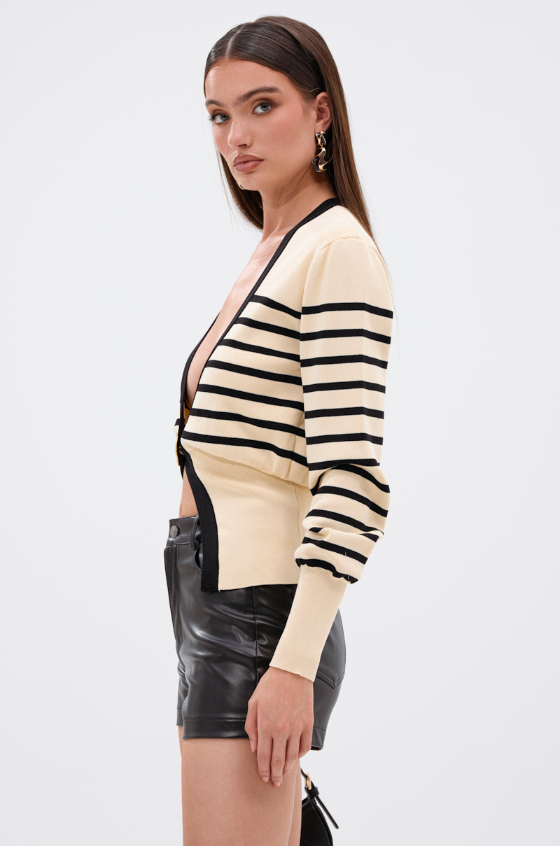 A woman with long brown hair stands in profile against a plain white background, wearing the REAL LIFE GOALS SWEATER—a cream and black striped knit cardigan with gold-tone buttons—paired with black leather shorts. 3