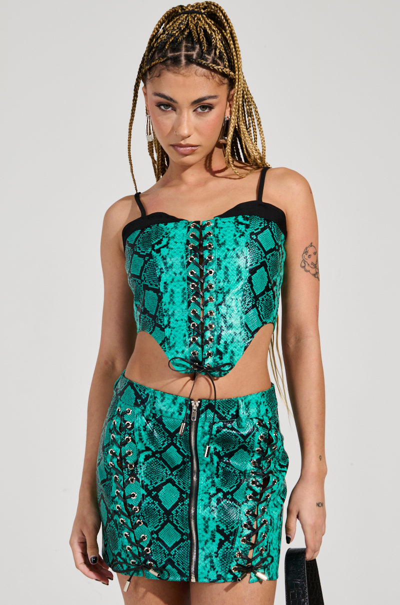 A person with braided hair is dressed in a turquoise faux snakeskin ensemble, highlighting the FOREVER THAT GIRL CORSET and a zip-up skirt. They have an arm tattoo and are posed against a plain background, holding a black handbag.