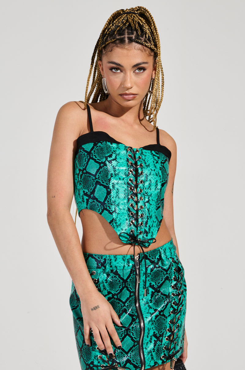 A person with braided hair wears the FOREVER THAT GIRL CORSET, a vibrant faux snakeskin ensemble in turquoise, featuring both a corset top and matching skirt. The outfit includes striking black lace-up details, set against a plain background that enhances the stylish apparel.