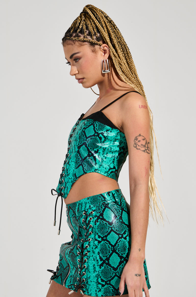 Dressed in the FOREVER THAT GIRL CORSET with its faux snakeskin pattern, turquoise side cutouts, and corset-style lace-up details, a person with long braided hair stands proudly. Tattoos accentuate their arms while hoop earrings add flair against the neutral background.