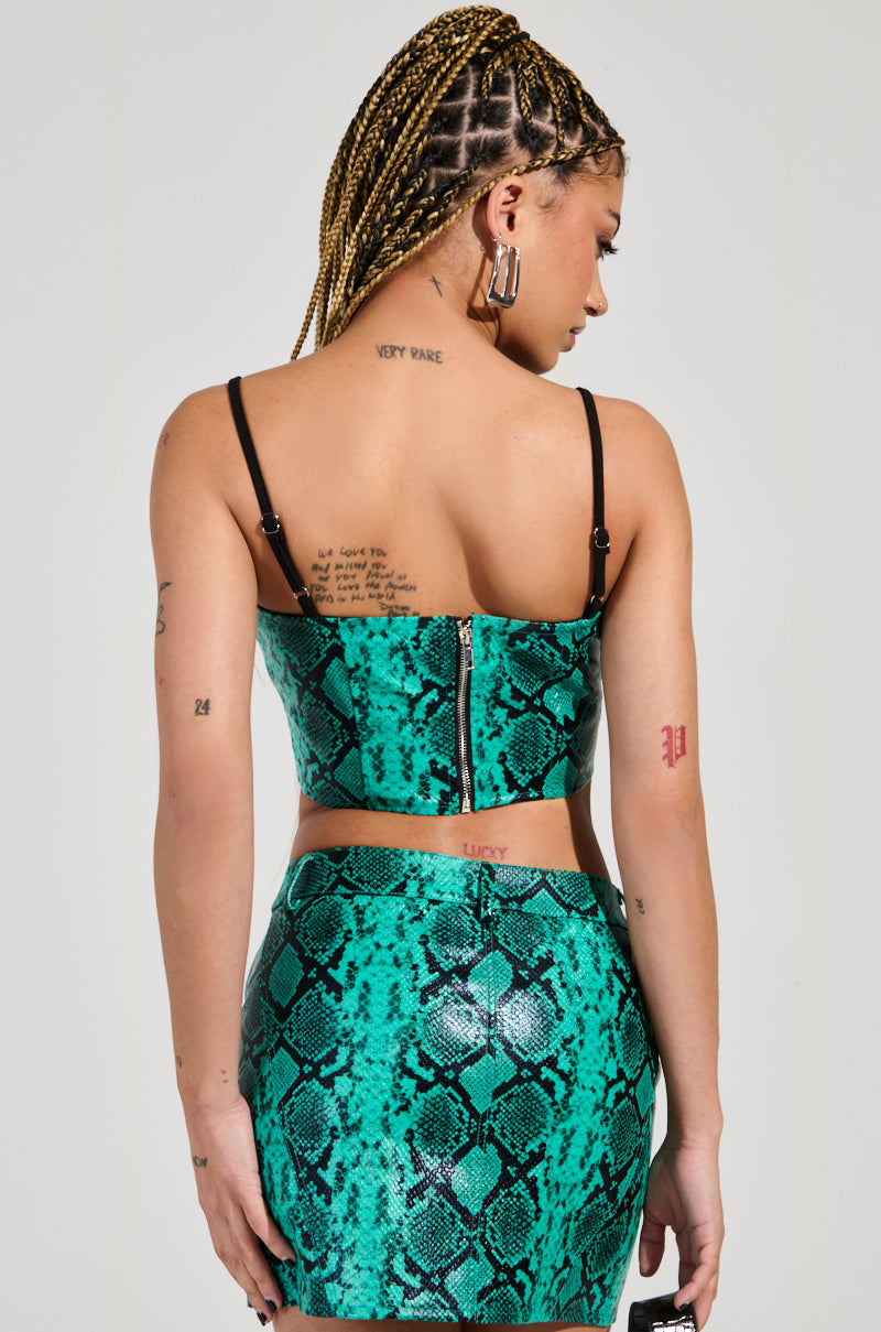A woman with braided hair wears the FOREVER THAT GIRL CORSET in a green and black animal print, along with a complementary skirt that features hints of faux snakeskin. Tattoos cover her back and arms as she holds a small black object in her left hand.