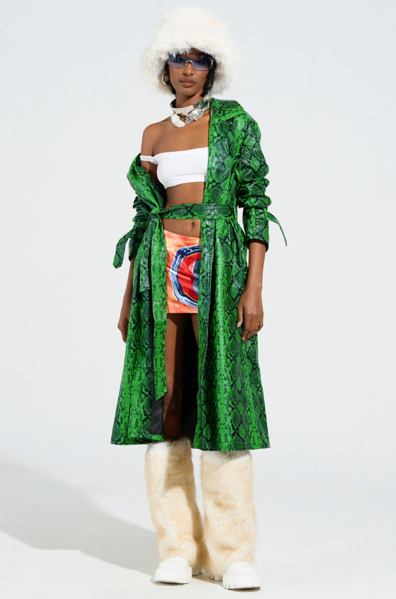 A person showcases a daring ensemble that includes the ENVY SNAKE PRINT TRENCH, paired with a white furry hat, sunglasses, a white tube top, colorful shorts, and white furry boots. The outfit is strikingly eclectic, with the embossed snakeskin print making a bold statement against the interplay of textures and colors.