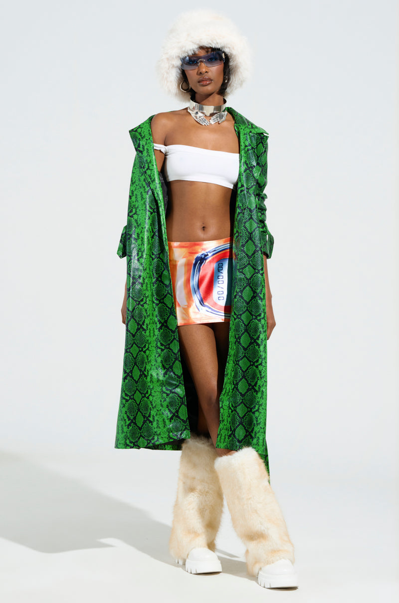Dressed in the ENVY SNAKE PRINT TRENCH, a fashion model radiates confidence, pairing it elegantly with a white crop top and a vibrant mini skirt. She accentuates her outfit with beige fluffy boots, a white furry hat, and sunglasses. A bold necklace adds the finishing touch to this chic ensemble set against a simple backdrop.