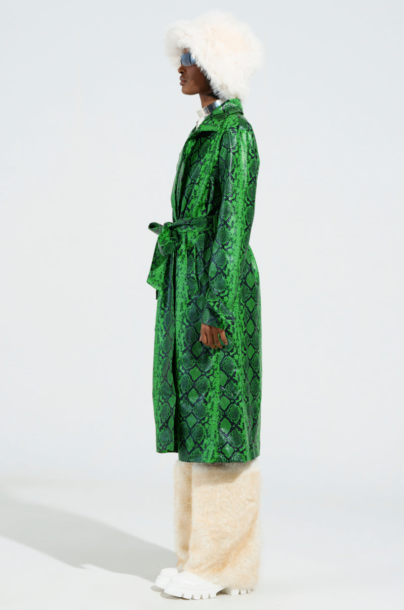 Against a plain background, a person stands in profile wearing the ENVY SNAKE PRINT TRENCH, complemented by a white fluffy hat, beige furry pants, and white boots. The coat's embossed snakeskin pattern enhances the boldness of the ensemble.