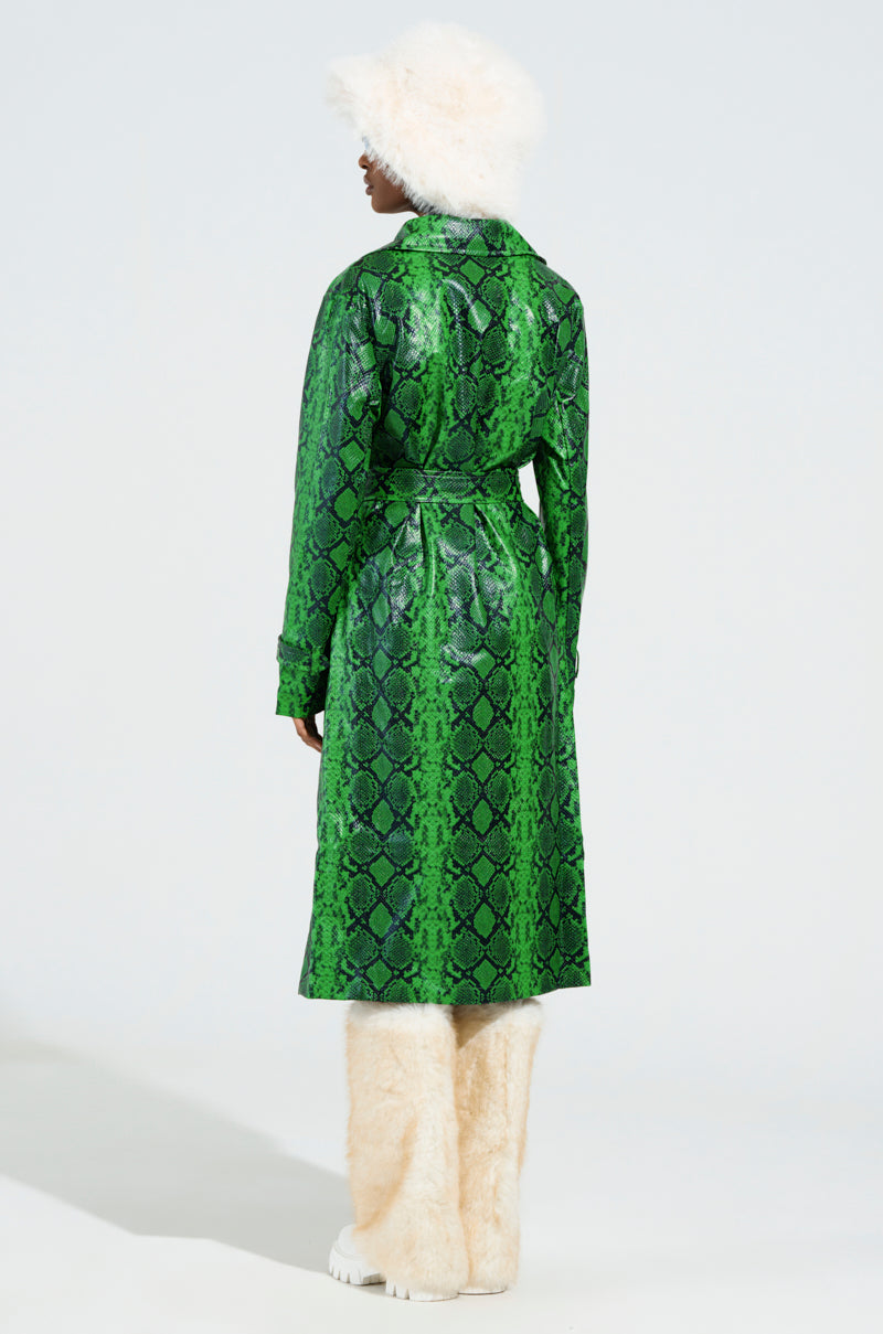 Dressed in the ENVY SNAKE PRINT TRENCH, a person pairs it with white furry boots and a matching white furry hat, creating a chic ensemble against a light, solid background.