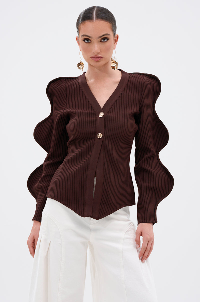 A woman wears the DON'T ASK CARDIGAN—a ribbed stretch knit in dark brown with sculptural wave sleeves and gold buttons—paired with high-waisted white pants, slicked-back hair, bold eye makeup, and large gold earrings. 0
