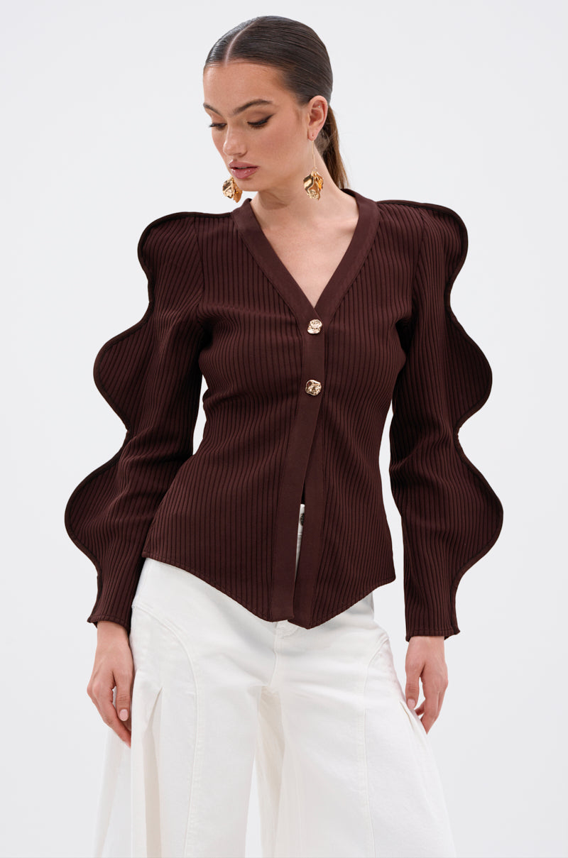 A woman poses against a plain white background wearing the DON'T ASK CARDIGAN—a ribbed stretch knit with sculptural wave-sleeve details and gold buttons—paired with high-waisted, wide-leg white pants and gold statement earrings. 1