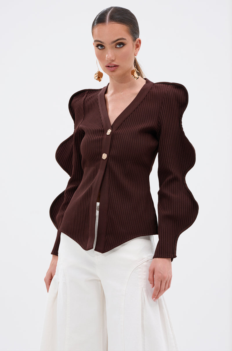 A woman models the DON'T ASK CARDIGAN in dark brown ribbed stretch knit with sculptural wave-sleeves and gold buttons, styled with high-waisted white pants, bold earrings, and slicked-back hair against a plain white background. 2