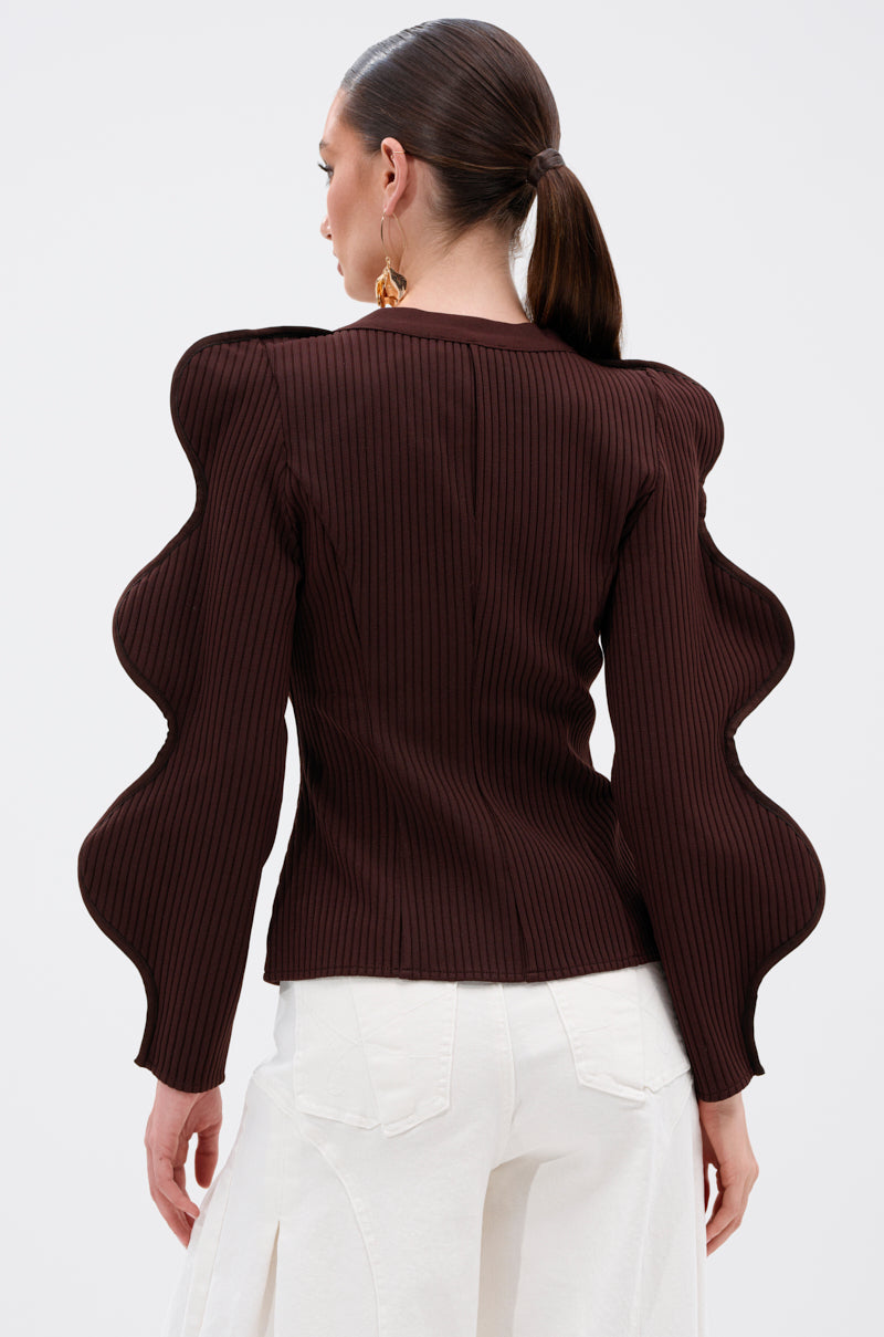 A woman with a low ponytail models the DON'T ASK CARDIGAN, a ribbed stretch knit top with sculptural wave sleeves. She faces away, paired with high-waisted white pants against a plain, light background. 4