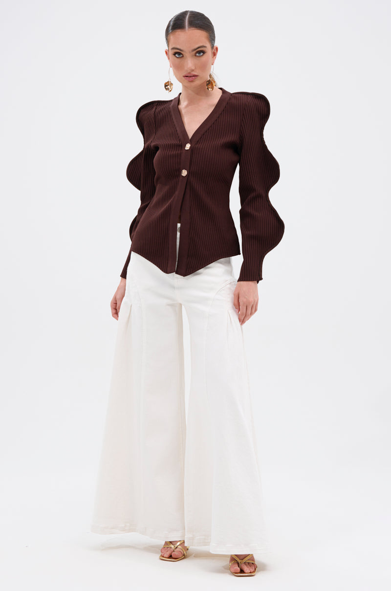 A woman models the DON'T ASK CARDIGAN, a dark brown ribbed stretch knit with sculptural wave-sleeve details and gold buttons, paired with high-waisted white wide-leg pants and gold open-toe heels against a white backdrop. 5