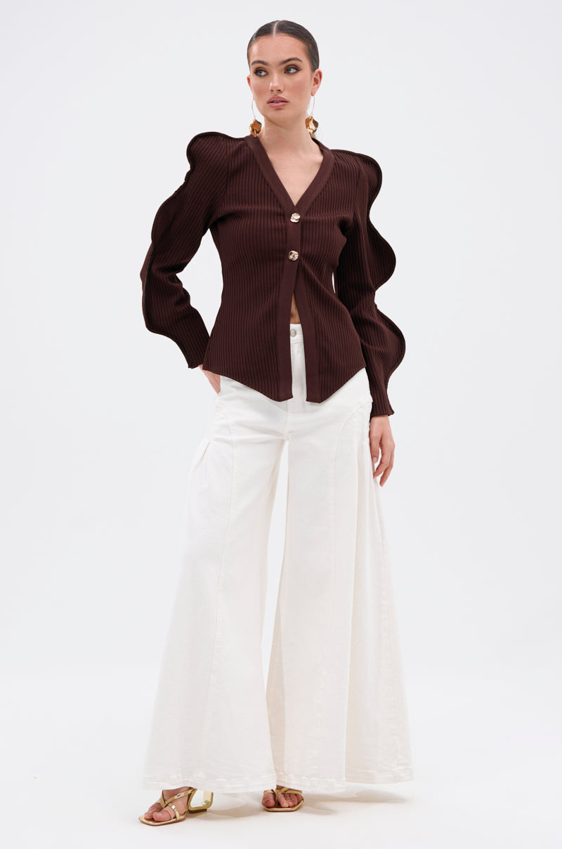 A woman poses in the DON'T ASK CARDIGAN—a ribbed stretch knit blouse with sculptural wave sleeves and gold buttons—paired with high-waisted, wide-leg white pants and gold flat sandals against a plain white background. 6