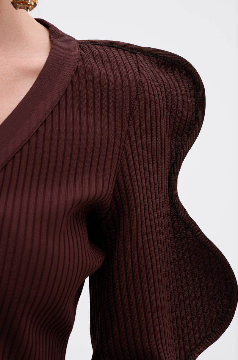Close-up of a person wearing the DON'T ASK CARDIGAN—a dark brown, ribbed stretch knit blouse with a V-neckline and sculptural wave-sleeve detail—against a light background. 7