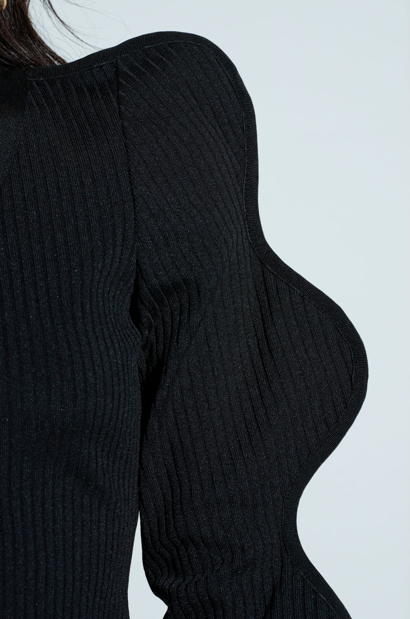 Close-up of a person wearing the WAVY CARDIGAN, showcasing its ribbed knit texture and voluminous sleeves. The high-fashion design silhouette stands out against the plain background.