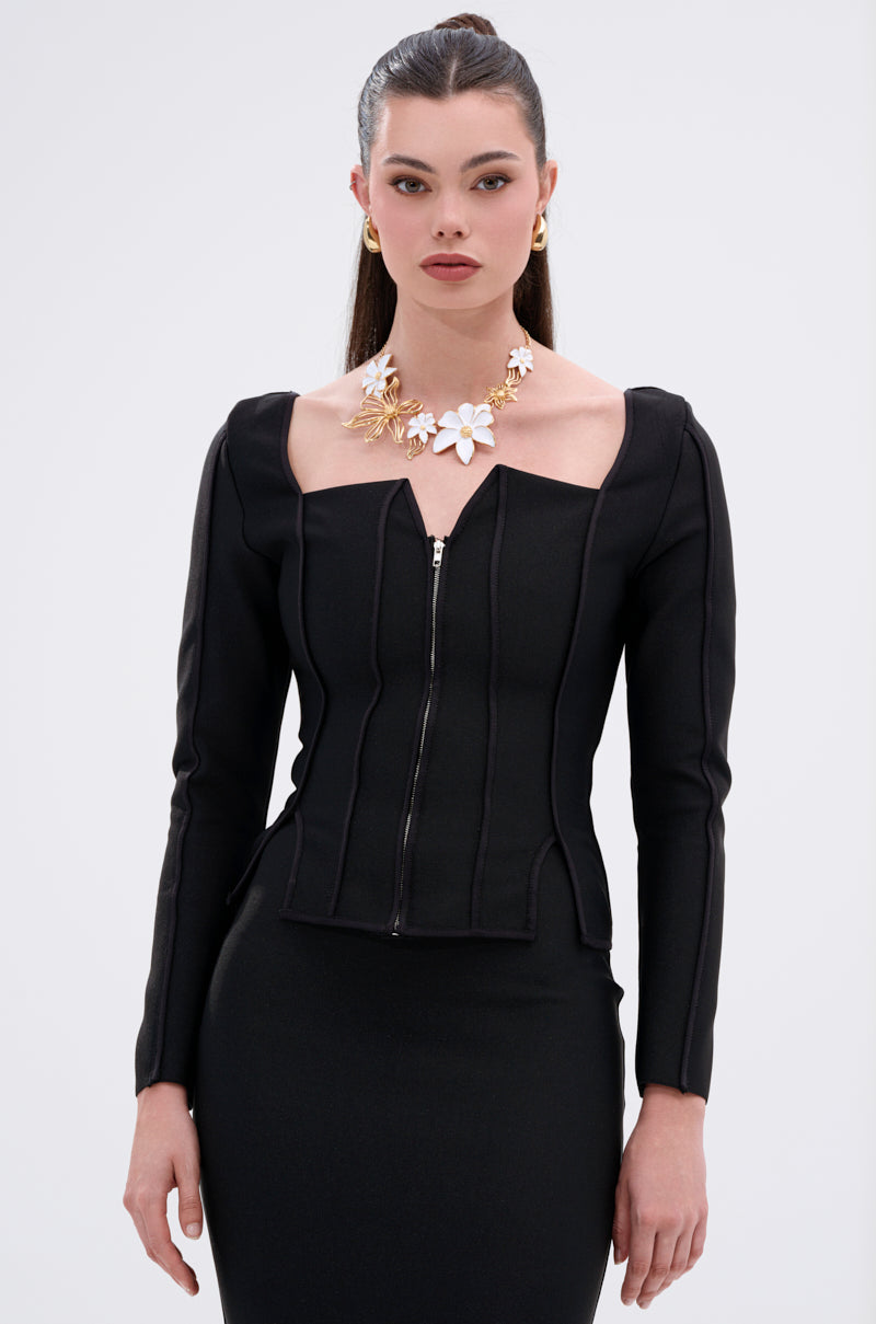 A woman with long dark hair styled back wears the JESSIE BANDAGE ZIP UP BLOUSE, a fitted black top with a corset-inspired bodice and metallic zip-front closure, accessorized by a floral statement necklace and gold earrings.