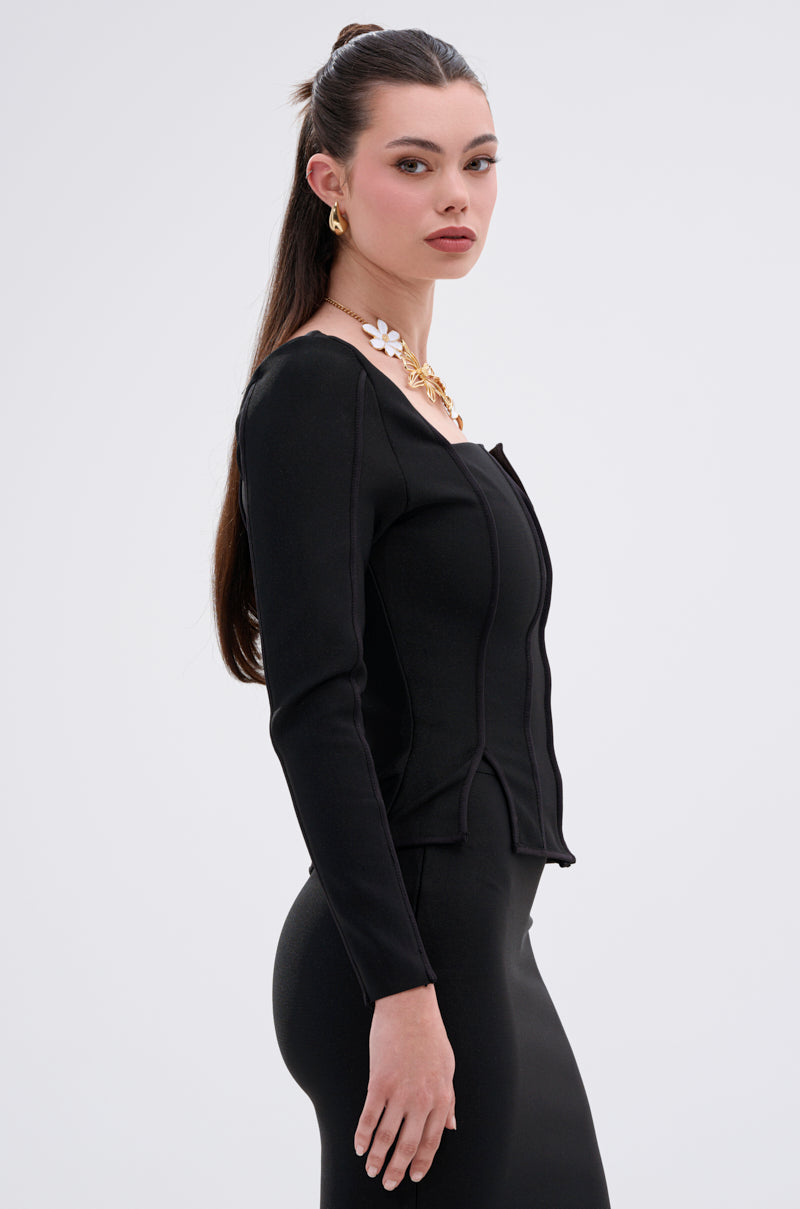 A woman with long, straight brown hair wears the fitted JESSIE BANDAGE ZIP UP BLOUSE and skirt, paired with gold earrings and a gold necklace with white flower accents. She stands in profile against a plain light background.