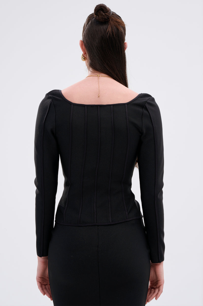 A woman with long brown hair, styled half-up, models the fitted JESSIE BANDAGE ZIP UP BLOUSE in black with vertical seams and a square neckline. She faces away from the camera, highlighting the back against a plain background.