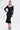 A woman poses in the JESSIE BANDAGE ZIP UP BLOUSE, a fitted black top with a metallic front zipper, paired with a knee-length mermaid skirt. She accessorizes with black heels and a floral necklace, her hair styled in a sleek ponytail.