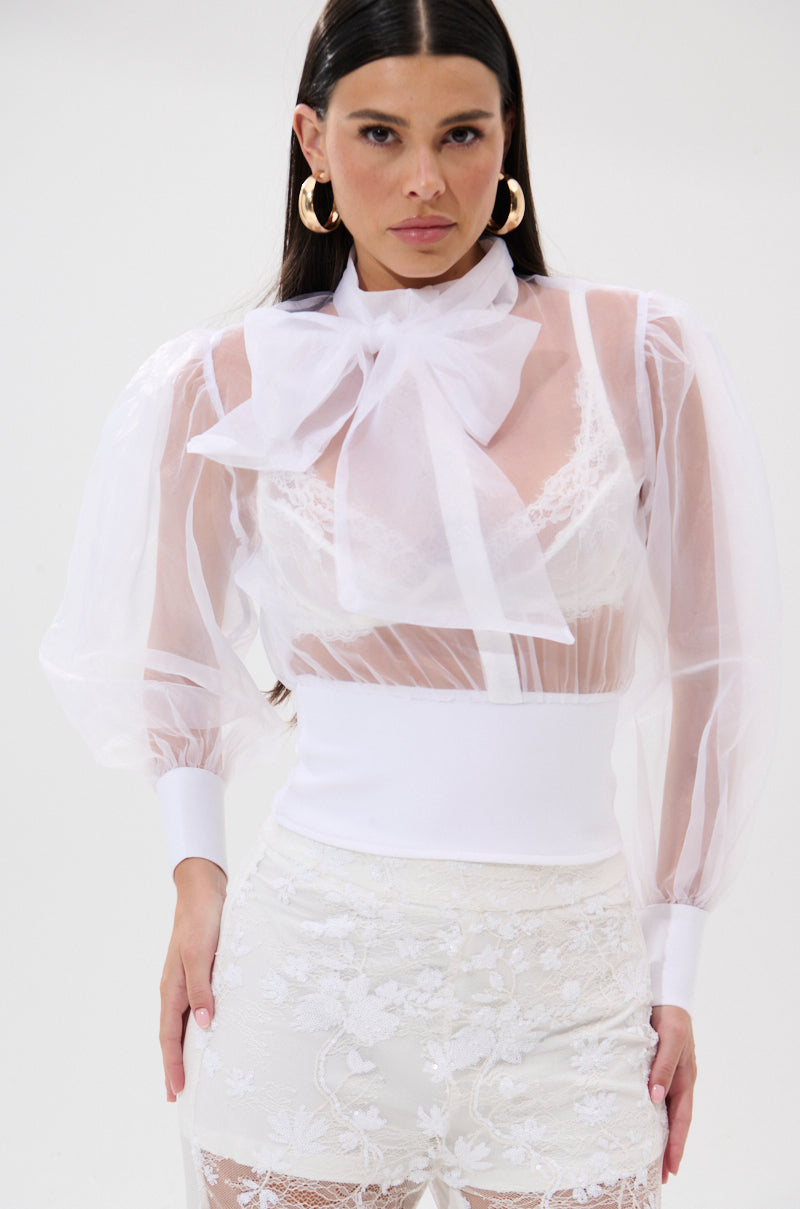 A woman wears the DANCE AND SHAKE BLOUSE WITH BOW IN WHITE, featuring sheer fabric, puffy sleeves, and a large neck bow. She pairs it with high-waisted lace pants, accessorizes with gold hoop earrings, and stands against a plain light background. 0
