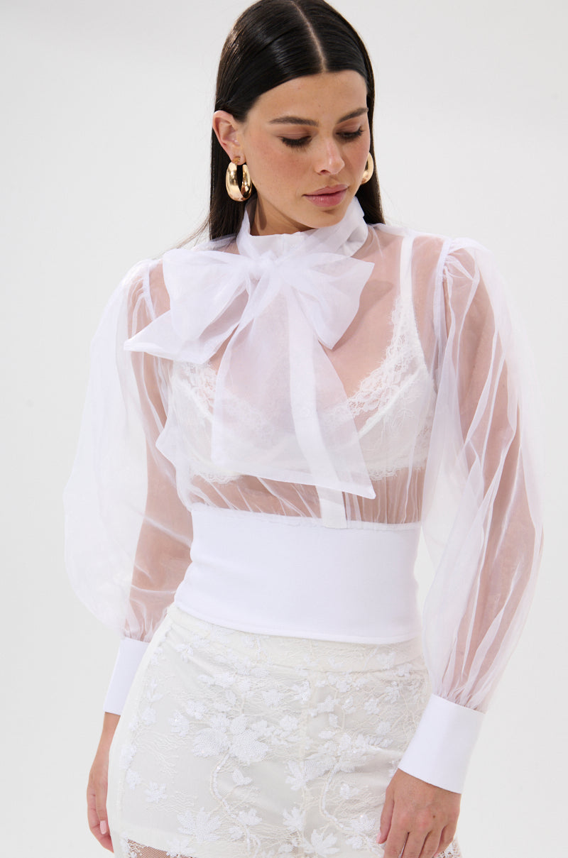 A woman wears the DANCE AND SHAKE BLOUSE WITH BOW IN WHITE, featuring puffy long sleeves and white cuffs, layered over a lace bralette. She styles it with a white embroidered skirt and gold hoop earrings for a chic statement look. 1