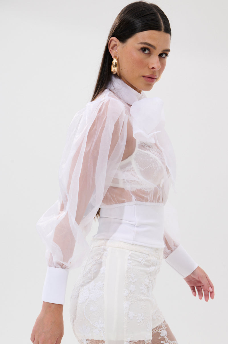 A woman with straight dark hair models the DANCE AND SHAKE BLOUSE WITH BOW IN WHITE, featuring sheer fabric and puffed sleeves. She pairs it with white lace shorts and faces sideways toward the camera against a plain background. 2