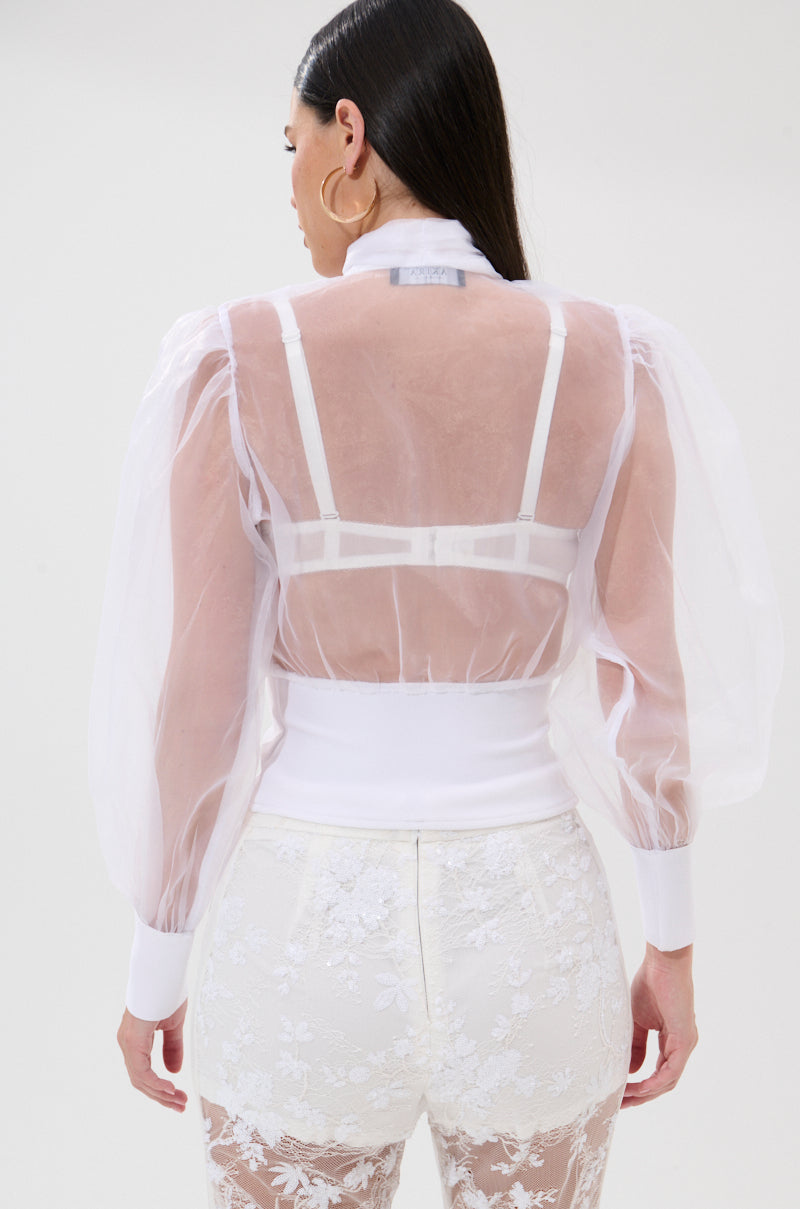 A woman with long dark hair is seen from behind, wearing the DANCE AND SHAKE BLOUSE WITH BOW IN WHITE—a sheer, airy puff-sleeve top over a white bra, paired with high-waisted floral lace shorts against a plain light background. 3