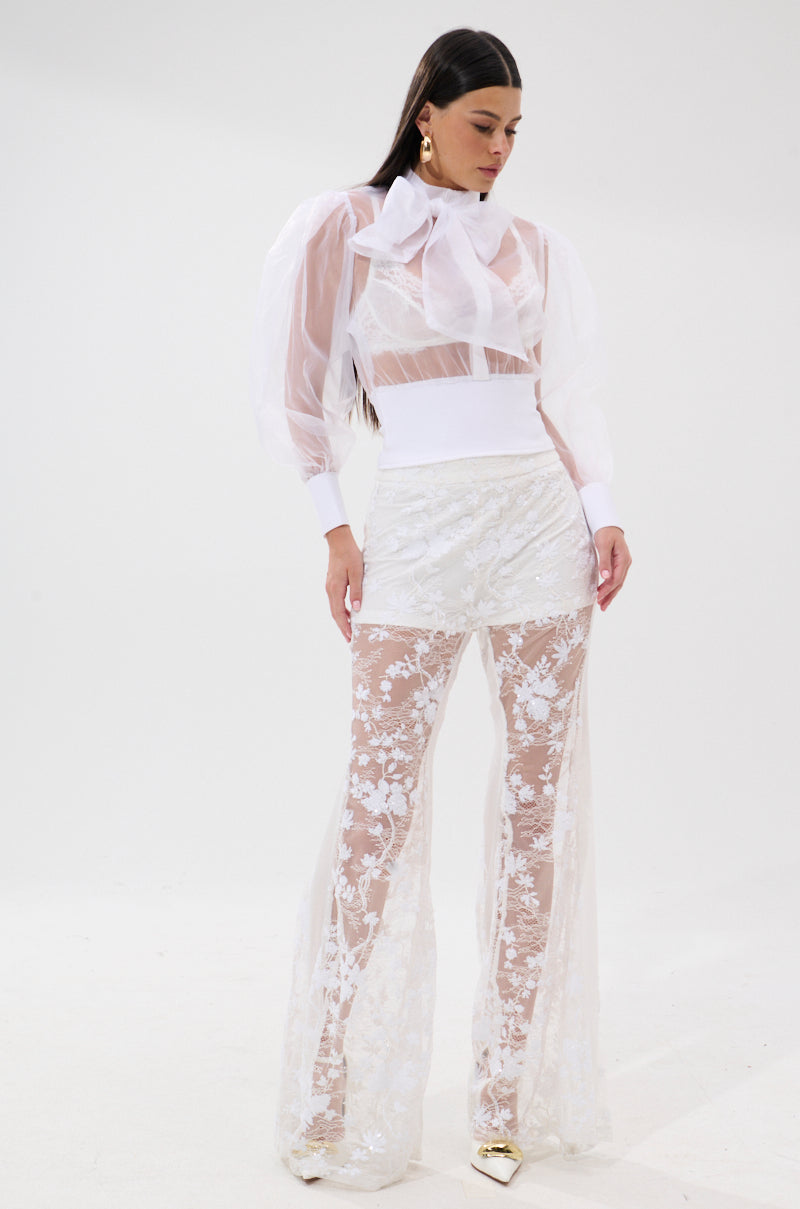 A woman with long dark hair stands against a white background wearing the DANCE AND SHAKE BLOUSE WITH BOW IN WHITE, paired with high-waisted sheer lace pants over matching shorts and gold pointed heels. 4