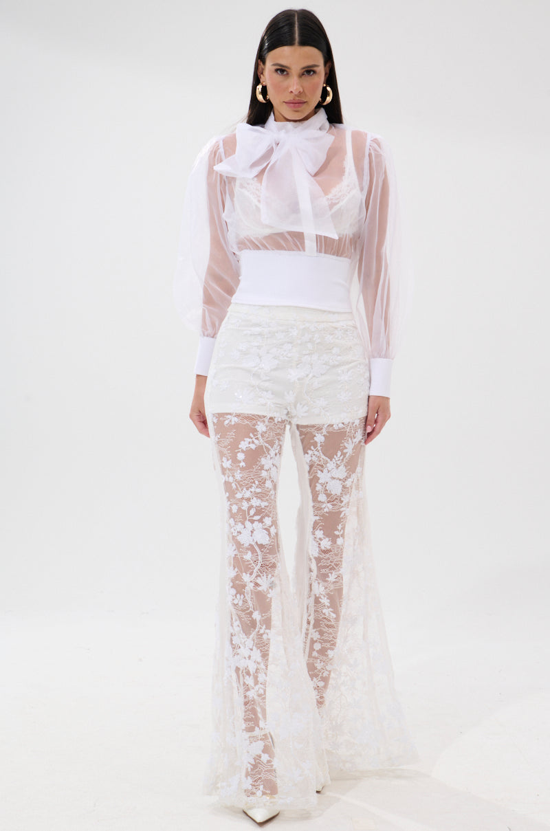 A woman with long dark hair and hoop earrings stands against a white background wearing the DANCE AND SHAKE BLOUSE WITH BOW IN WHITE, a sheer blouse featuring a large neck bow, paired with high-waisted, flared floral lace pants. 5