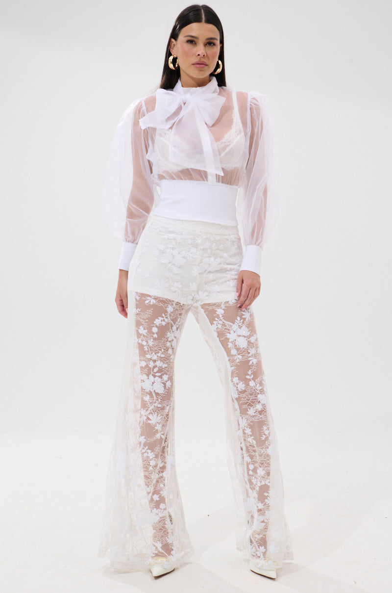 A woman poses against a white backdrop in the DANCE AND SHAKE BLOUSE WITH BOW IN WHITE, styled with wide lace flare pants over shorts, white pointed heels, and gold hoop earrings. 6